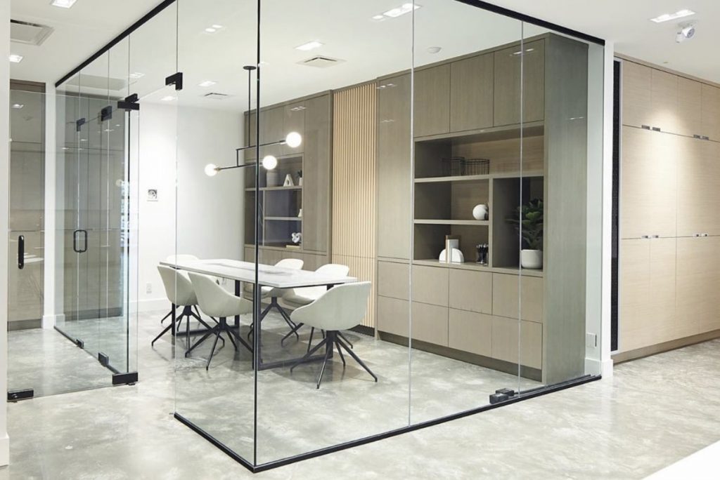 A modern conference room with glass partitions, a long white table, and several white chairs. The room has minimalist decor, with built-in wooden shelving units and a contemporary light fixture hanging above the table. The floor is made of polished concrete.