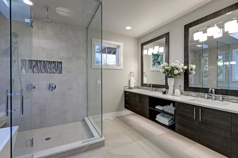 A modern bathroom with a large, glass-enclosed shower on the left, featuring dual shower heads and glass walls. The right side has a double-sink vanity with dark wood cabinets, a marble countertop, and a large mirror. The room is well-lit, with a bouquet of white flowers on the counter.