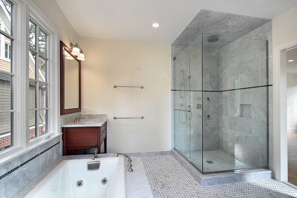 A modern bathroom featuring glass walls around a shower with marble tiles, a soaking bathtub with jets, and a wooden vanity with a large mirror. The room is well-lit by natural light from a large window beside the bathtub. The walls are painted light beige.