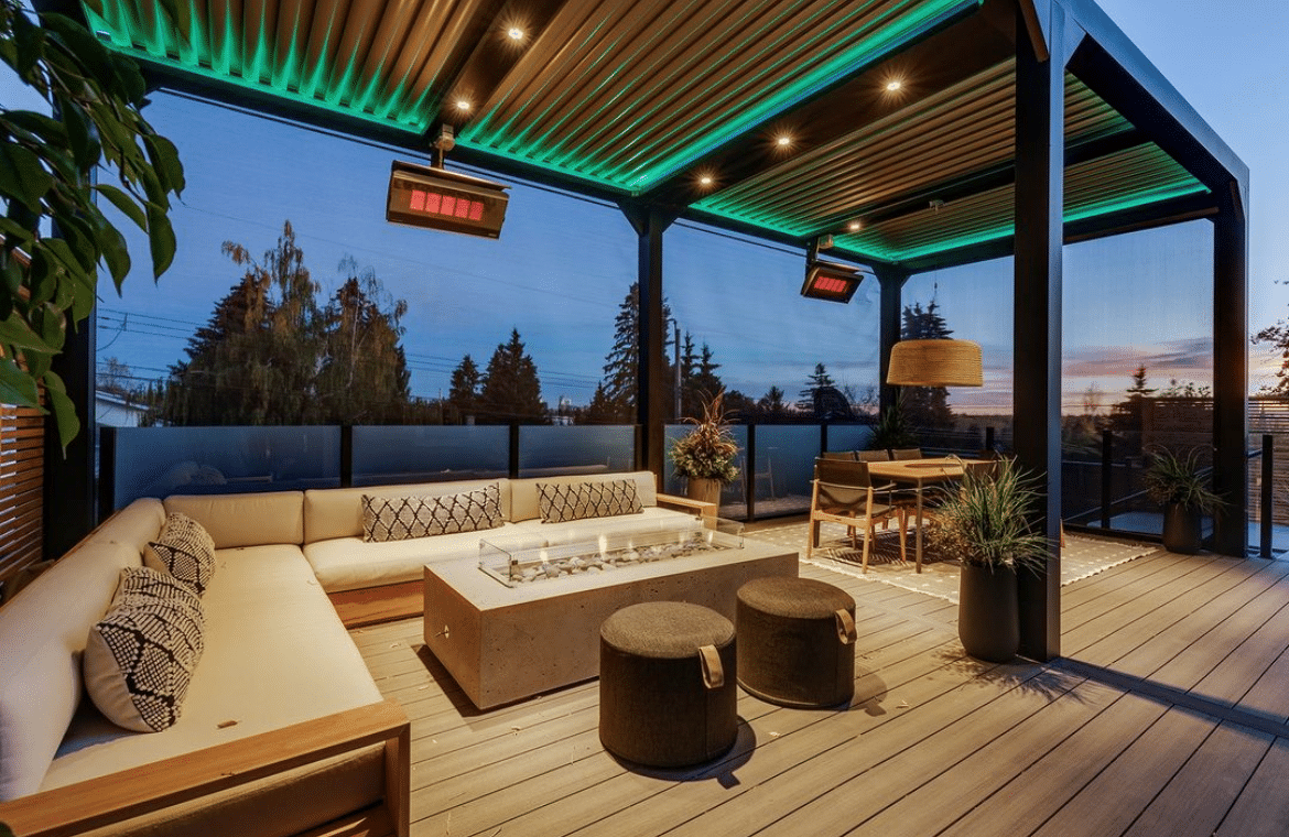 A modern outdoor patio during dusk features a stylish seating area with an L-shaped couch, a concrete fire pit, and two black ottomans. The space is illuminated by green overhead lights and complemented by plants, a dining table, glass walls, and a semi-transparent screen.