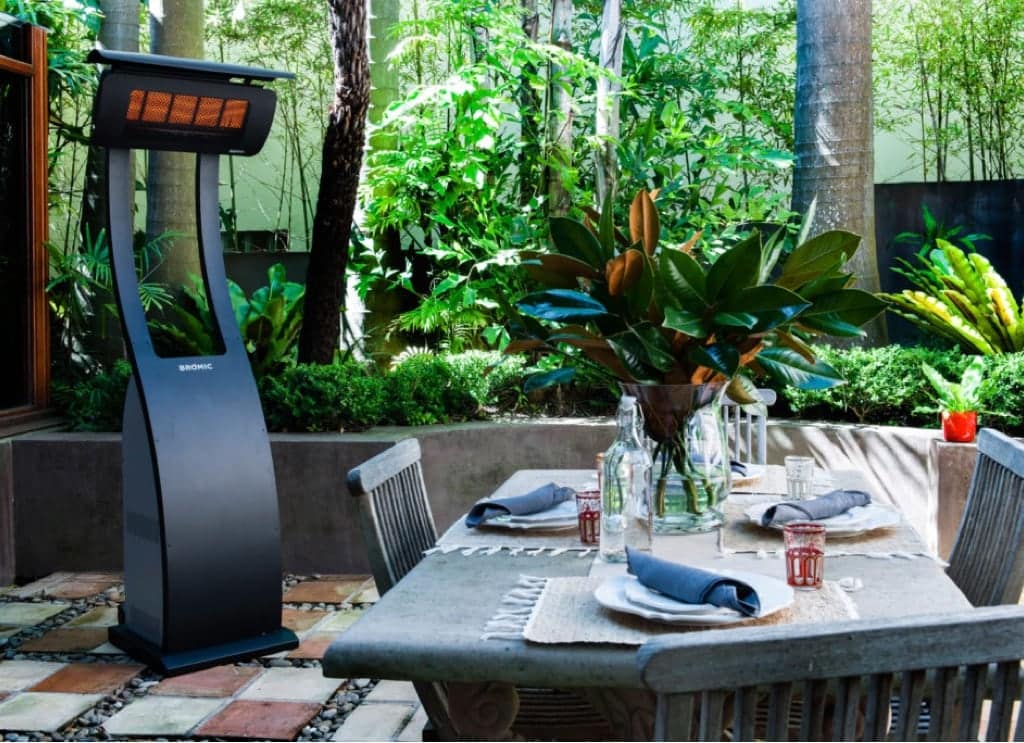 A well-lit outdoor dining area features a neatly set table with four place settings, each adorned with folded napkins. A standing patio heater to the left ensures warmth and comfort. The background, with its lush green plants and trees, creates a serene and inviting ambiance.