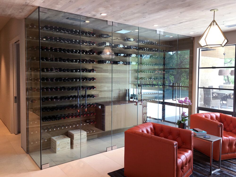 Modern wine cellar with glass walls, featuring numerous wine bottles displayed on wall-mounted racks. The room has wood panels, two red tufted armchairs, a table with reading materials, and soft lighting from a geometric pendant light. An outdoor garden is visible through the windows.