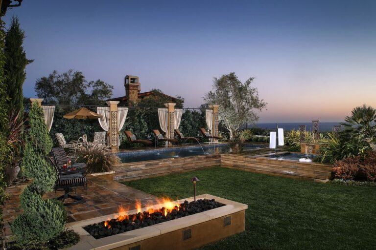 A luxurious backyard at dusk features a rectangular pool with lounge chairs and draped pergolas. Glass walls seamlessly blend indoor and outdoor spaces, enhancing the view of the stone fire pit with roaring flames, surrounded by manicured grass and lush landscaping. Trees and a house are visible in the background.