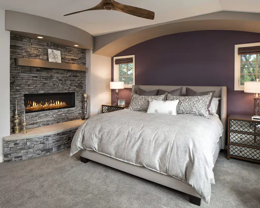 A modern bedroom featuring glass walls and a stone fireplace with a built-in electric fire, a king-sized bed with a taupe comforter and patterned pillows, and two matching nightstands with table lamps. The walls are painted purple and grey, and a wooden ceiling fan is overhead.