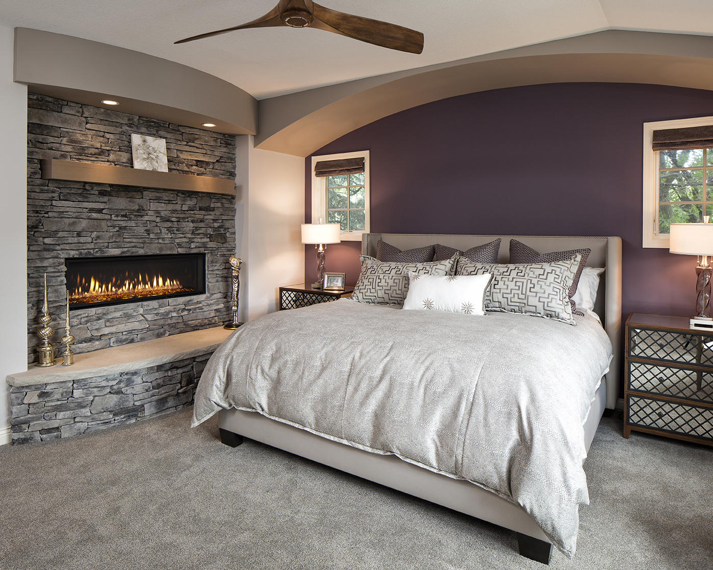 A modern bedroom featuring glass walls and a stone fireplace with a built-in electric fire, a king-sized bed with a taupe comforter and patterned pillows, and two matching nightstands with table lamps. The walls are painted purple and grey, and a wooden ceiling fan is overhead.