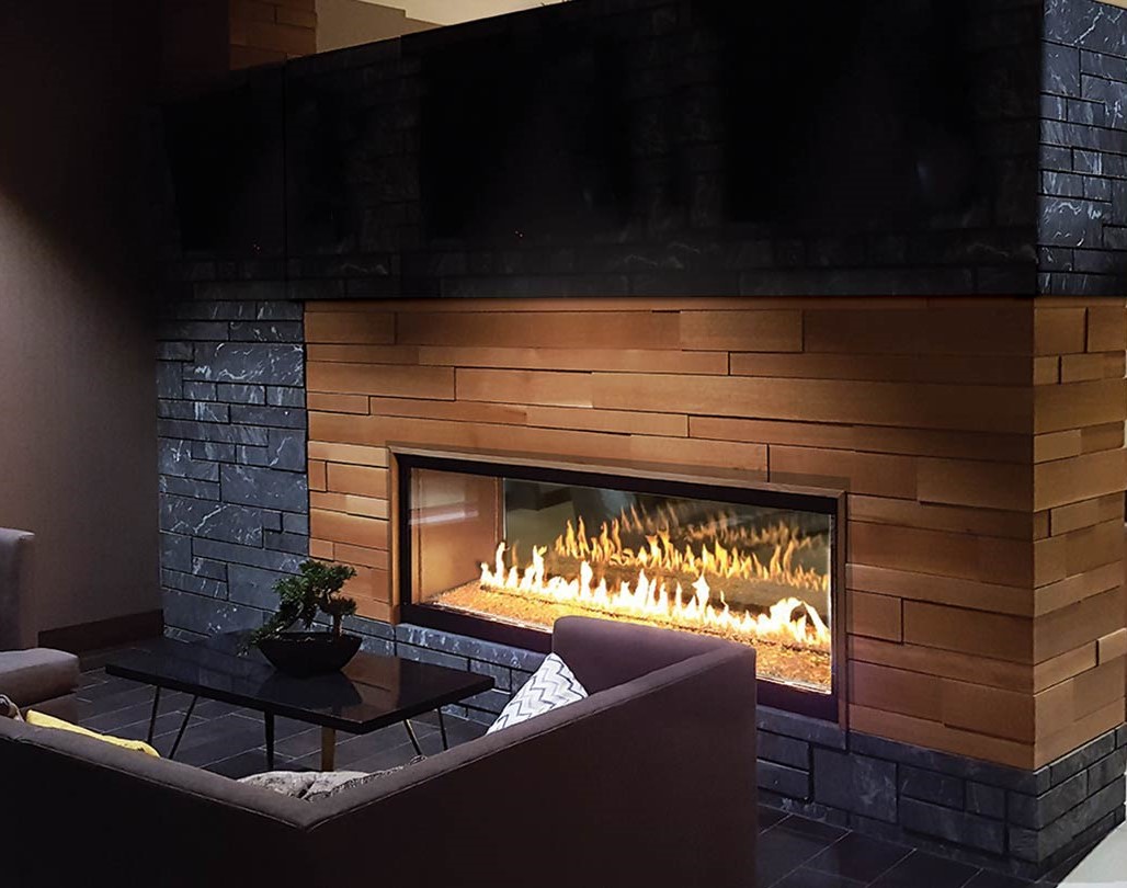 A modern living room features a sleek, large glass-enclosed fireplace set in a wall with wooden and dark stone accents, forming the stylish foundation of the space. In front of the fireplace, a gray sectional sofa and a small black coffee table with a plant create a cozy seating area.