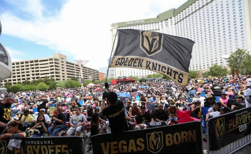 A large crowd gathered outdoors, many wearing black and gold attire representing the Vegas Golden Knights, holding team flags and banners. A man in the foreground waves a large “Golden Knights” flag. The Park MGM hotel is visible in the background. A banner reads "VEGAS BORN.