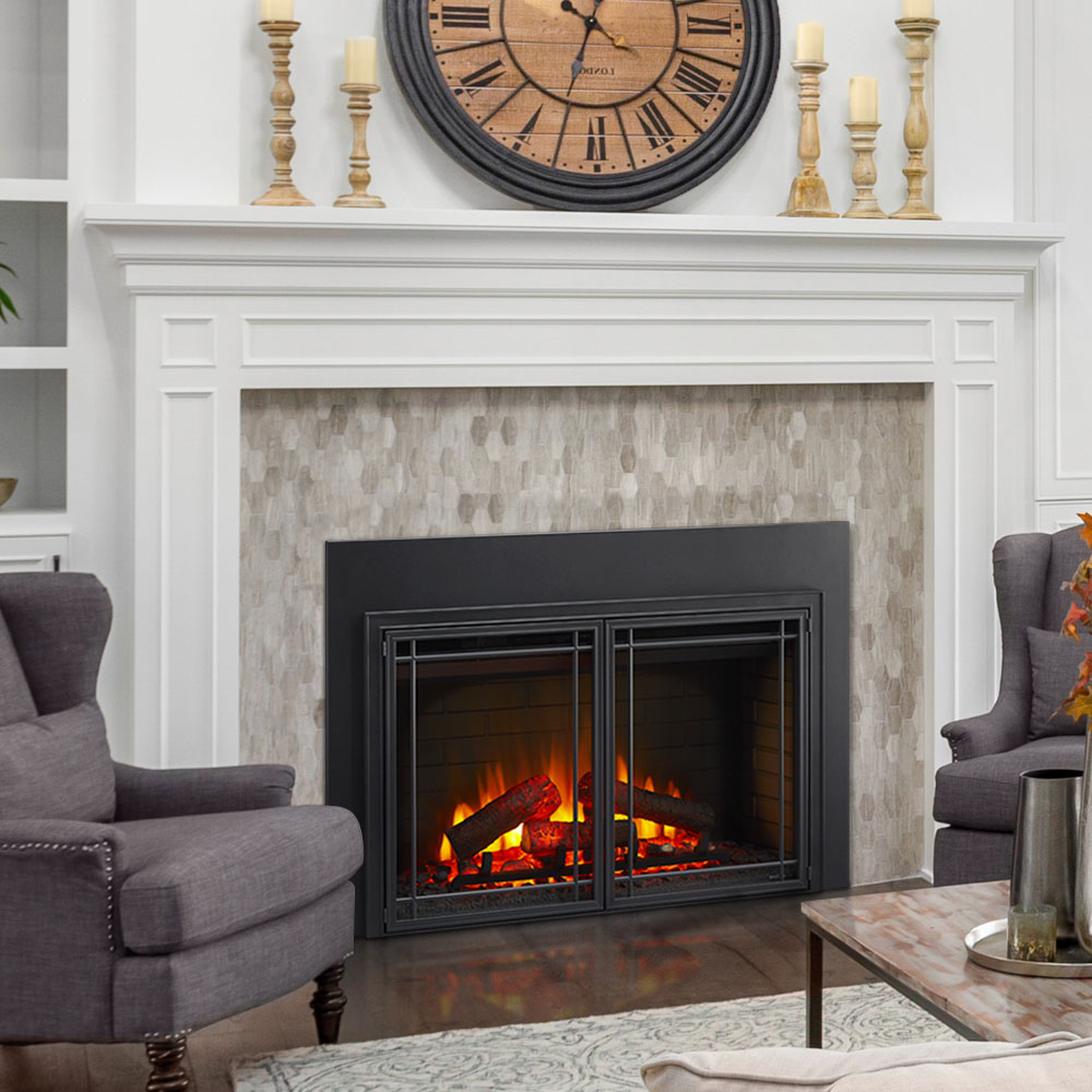 A cozy living room featuring a lit fireplace at the center with a black frame and white mantel. The mantel is adorned with candles and a large wooden clock. Two gray armchairs flank the fireplace, while glass walls allow natural light to flood in, highlighting the coffee table in front of the hearth.