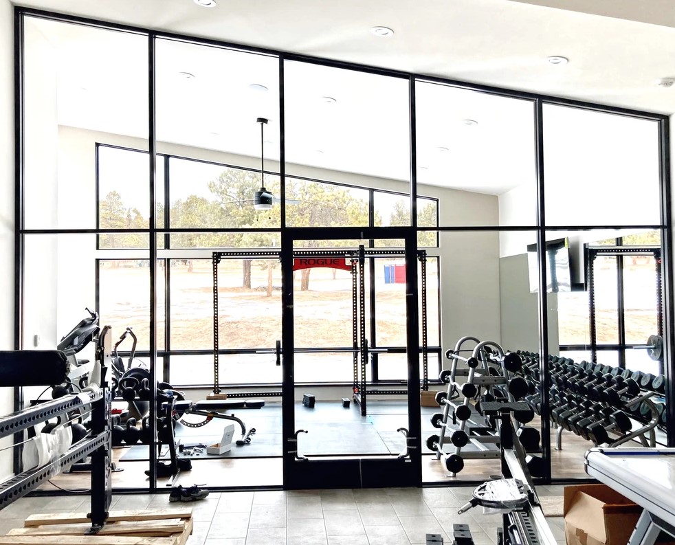 A modern gym with expansive glass walls letting in natural light. The gym includes various exercise equipment such as treadmills, weightlifting racks, a bench press, and a variety of free weights organized on racks. Outside, trees and a lawn are visible.