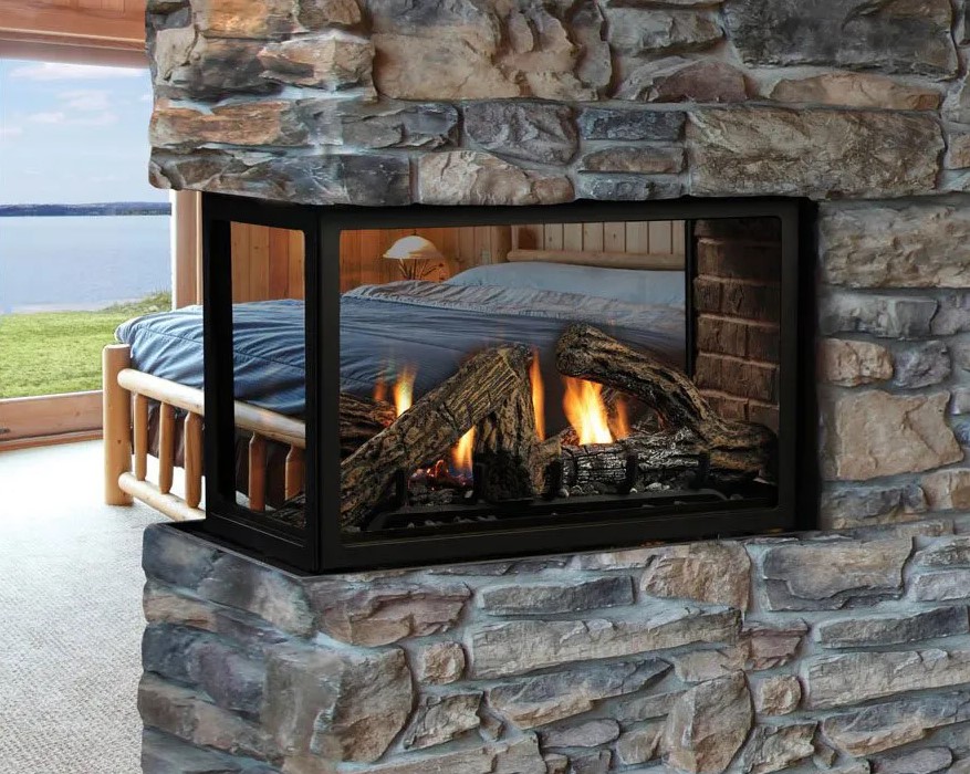 A room with a bed and a large window overlooking a body of water. In the foreground, there is Heatilator's Three-Sided Gas Fireplace with visible flames, providing a warm and cozy atmosphere. The wooden bed with blue bedding is positioned to enjoy the view.