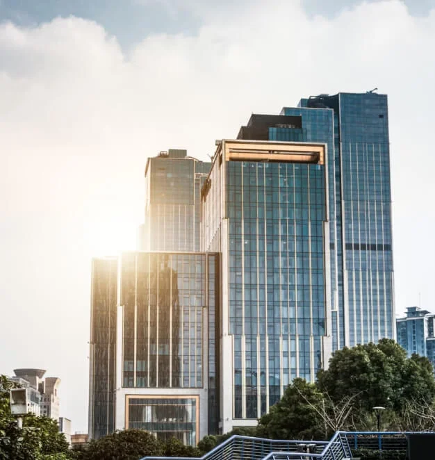 A group of modern high-rise office buildings with large glass windows reflecting sunlight, almost like a sidebar against the blue sky. The buildings are surrounded by greenery and other urban structures, creating a vibrant contrast between nature and concrete.