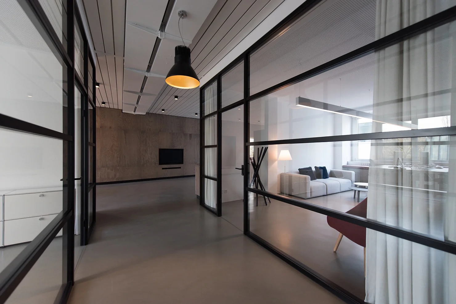 Modern interior with a minimalist design. Glass walls separate the hallway from the living area, which features a light gray couch, a floor lamp, and a TV mounted on a concrete wall. The space is well-lit with natural and artificial lighting.