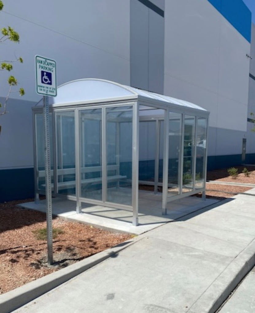 A modern bus stop shelter with transparent glass panels and a curved roof, situated on a cemented area next to a large building. Nearby, you'll find a pole with a "Handicap Parking" sign. The ground around the shelter is lined with mulch and small plants, making us feel welcome and inclusive.