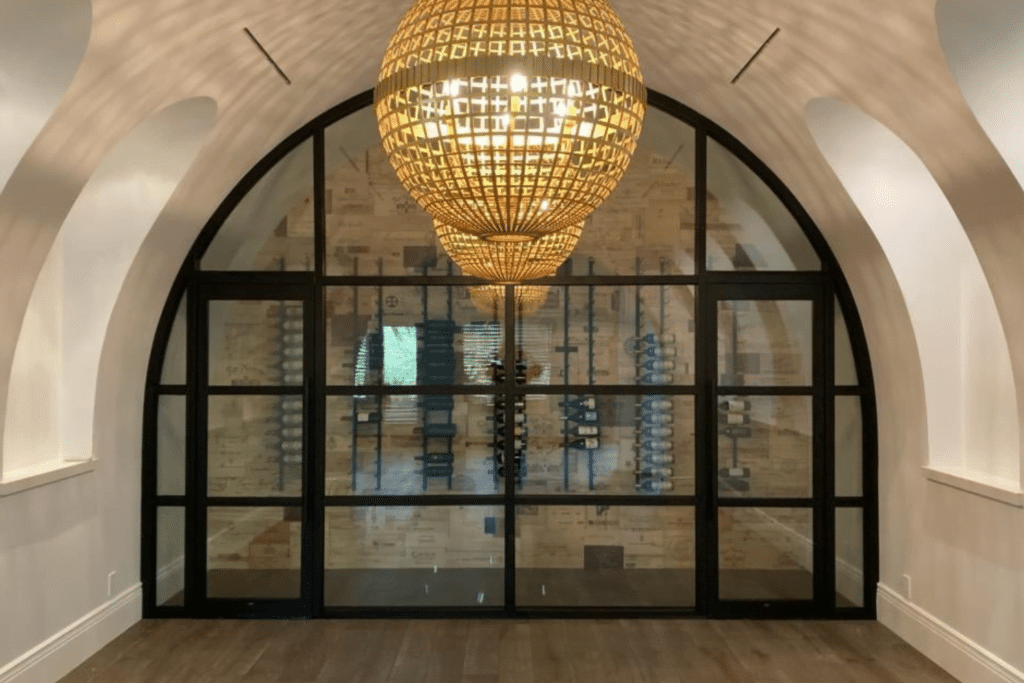 A luxurious wine cellar by Hearth & Home Specialties features a large, ornate chandelier and arched glass doors with black framing. The interior showcases wooden wine racks lining the walls and a neatly organized collection of wine bottles. The room has white walls and dark hardwood flooring.