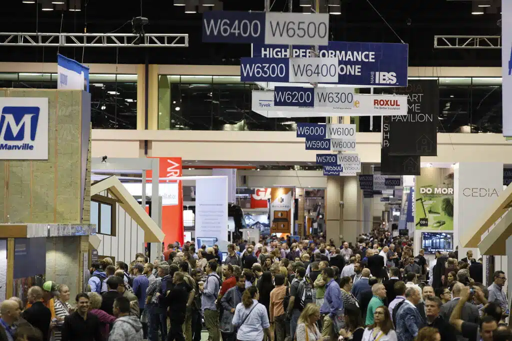 Crowd at a convention or trade show with booth signs hanging above and people walking around.