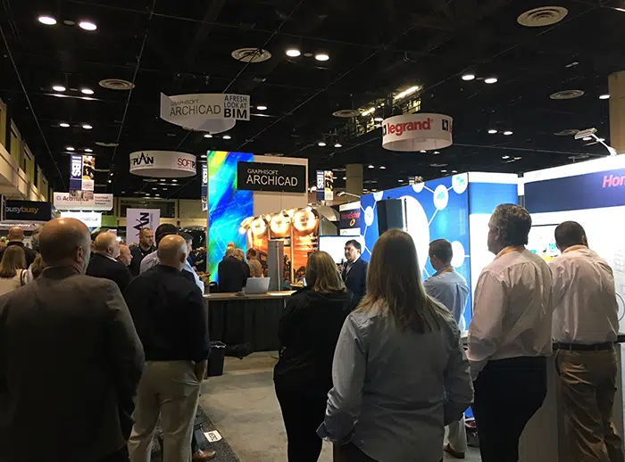 People attending a trade show, gathered around booths with banners overhead.