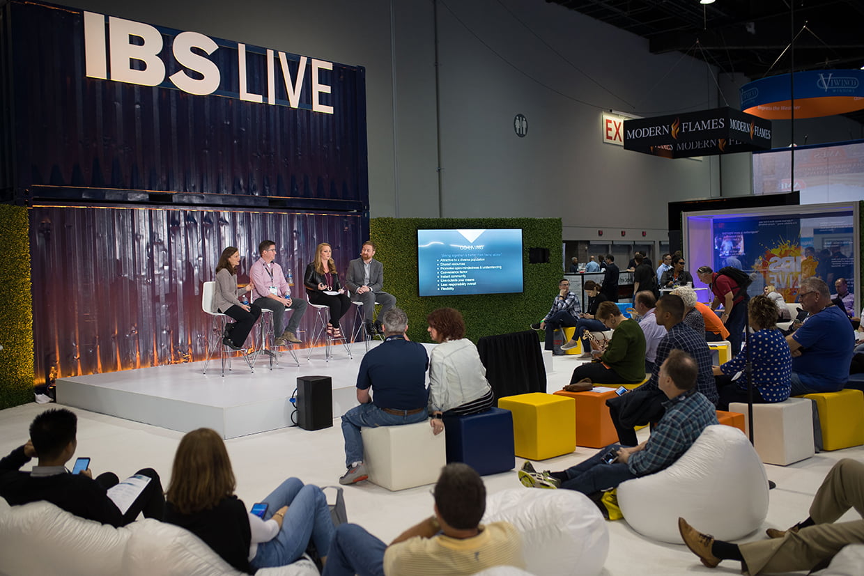 A panel of four people is seated on a stage under the sign "IBS LIVE," facing an attentive audience. The audience, comfortably settled on colorful cubes and white bean bags, watches intently as a screen beside the panel displays a presentation slide. The event unfolds in a spacious, well-lit venue.