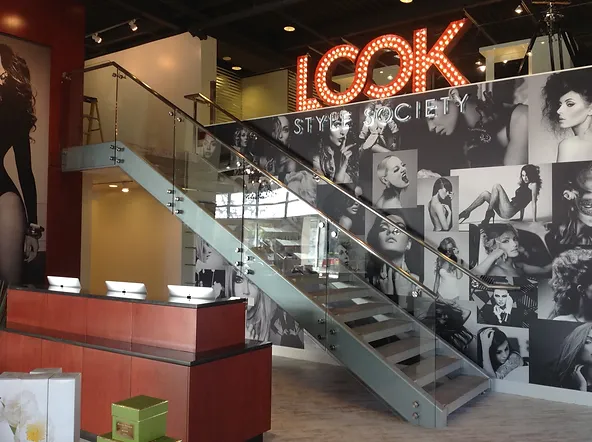 A modern, stylish interior of a salon features a prominent staircase with glass railings leading upstairs. The wall is adorned with large, black-and-white photos of various models. Glass walls enhance the airy atmosphere, and a large illuminated sign reads "LOOK Style Society." The reception desk is in the foreground.