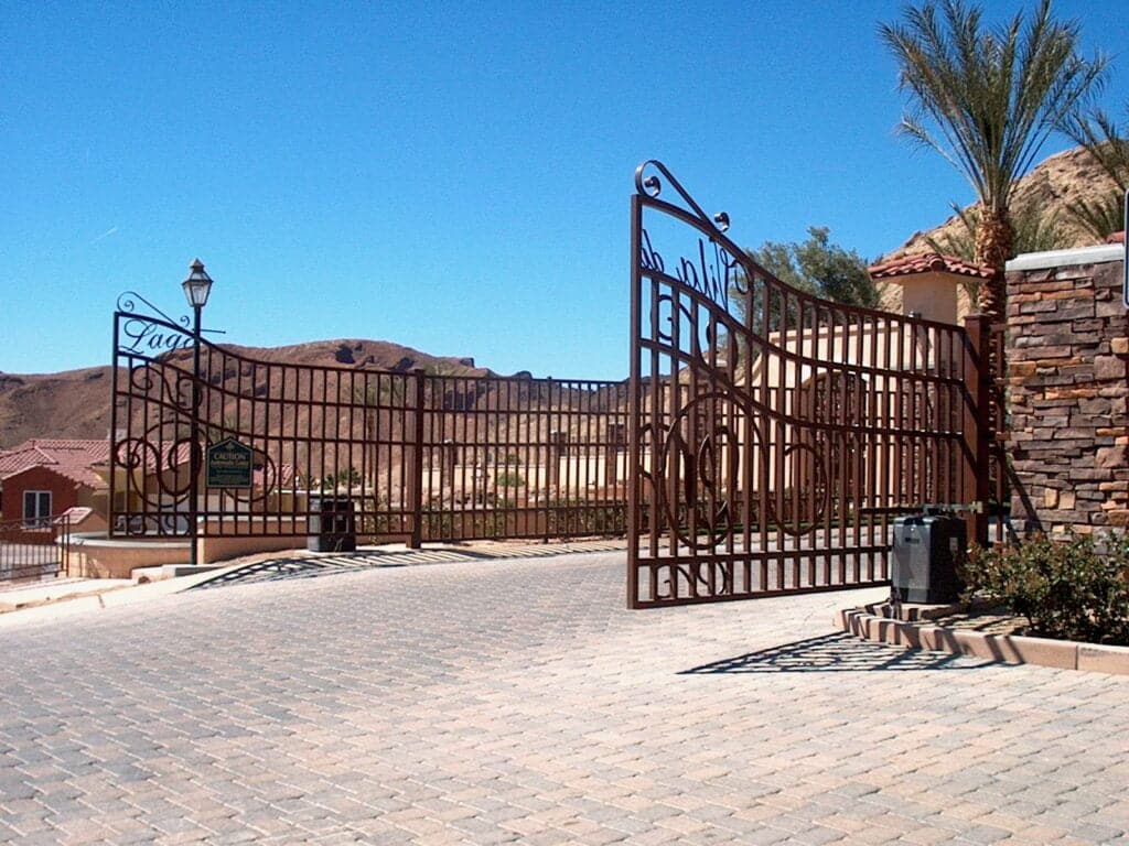 Photo of a grand, ornate wrought-iron vehicle entry gate at the entrance of a residential community. The gate is open, revealing a cobblestone pathway leading into the area, bordered by stone walls and palm trees. Hills are visible in the background under a clear blue sky.