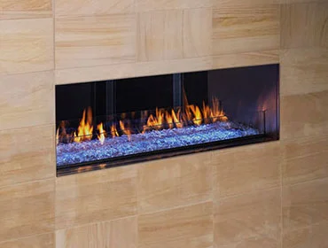 A modern, rectangular fireplace features a sleek design with vibrant flames dancing behind a glass panel. The surrounding wall, seamlessly integrated with glass walls, is covered in smooth, light-colored tile. The glass-enclosed fire burns on a bed of decorative crystals, creating a warm ambiance.