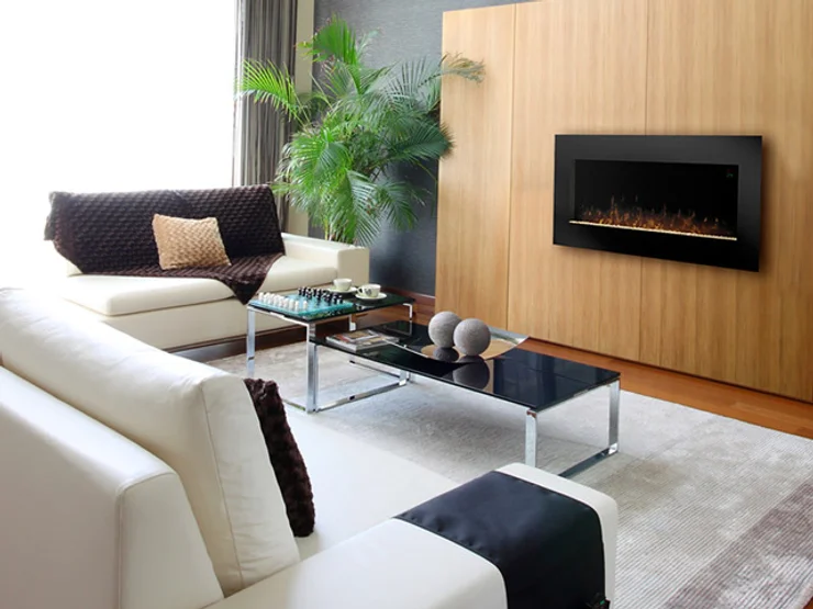 A modern living room with white sofas, a glass coffee table with decor items, a chess board, and a potted plant beside the sofa. The space features glass walls, enhancing natural light. A wall-mounted electric fireplace is on a wooden wall panel, adding warmth to the minimalist and cozy ambience.