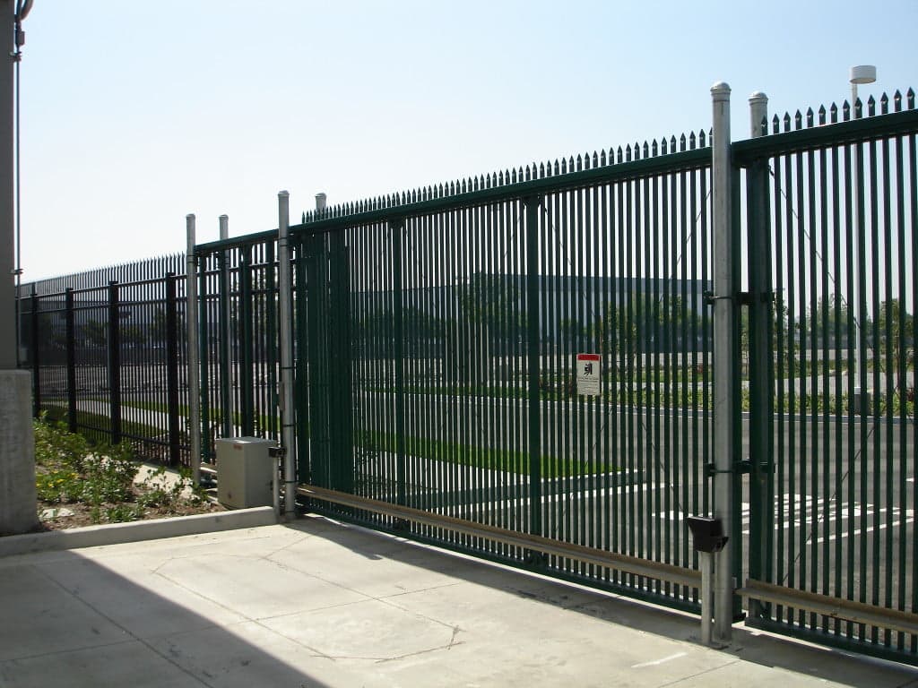 A tall metal fence with narrow bars and a sliding gate partially open marks the entry to a large paved area, where some greenery is visible. A small sign is attached to the fence, and a security box is mounted on the side. Industrial buildings loom in the background, hinting at vehicle access control.