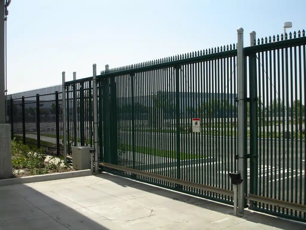 A tall metal fence with narrow bars and a sliding gate partially open marks the entry to a large paved area, where some greenery is visible. A small sign is attached to the fence, and a security box is mounted on the side. Industrial buildings loom in the background, hinting at vehicle access control.