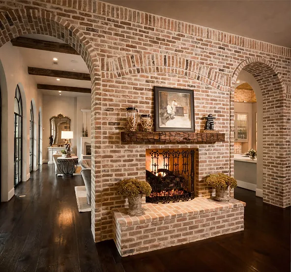 A cozy living space with a rustic brick archway and fireplace features dark wood floors, a warm fire in the hearth, and decorative plants on either side. The elegant furnishings and artworks complement the glass walls, creating an inviting and sophisticated ambiance.