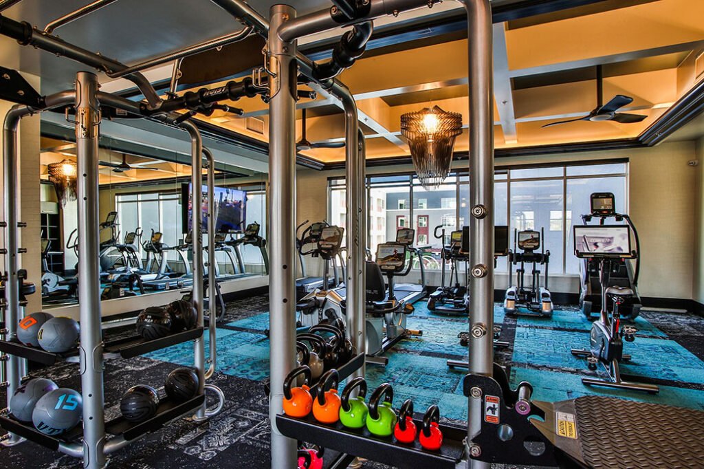 A modern gym with various exercise equipment, including dumbbells, kettlebells, a weight rack, stationary bikes, treadmills, and elliptical machines. Large mirrors cover one wall while windows provide natural light. The floor is carpeted in shades of blue and black. Convenient shower enclosures are available for post-workout refreshment.