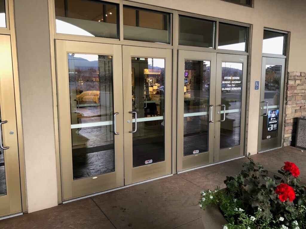 A glass entrance with four doors, each featuring horizontal push bars, framed in beige metal. Inside, a reflection shows indoor lighting and furnishings that resemble an inviting shower enclosure. A potted plant with red flowers and green leaves is visible in the foreground near the entrance.