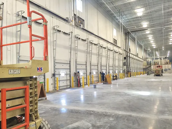 A large warehouse interior featuring several loading docks with closed doors. An elevated work platform is on the left, and some workers are involved in commercial door installation near the loading docks. The space is illuminated by overhead lighting.