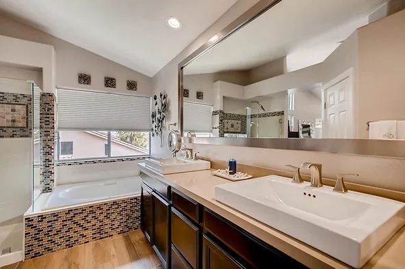 Modern bathroom with dual sinks and large mirror. Features a bathtub with mosaic tile backsplash, beige walls, and wooden floor. Glass walls allow natural light to flood the space. Ample counter space, a wide window with shades, and decorative accents enhance the room's aesthetics.