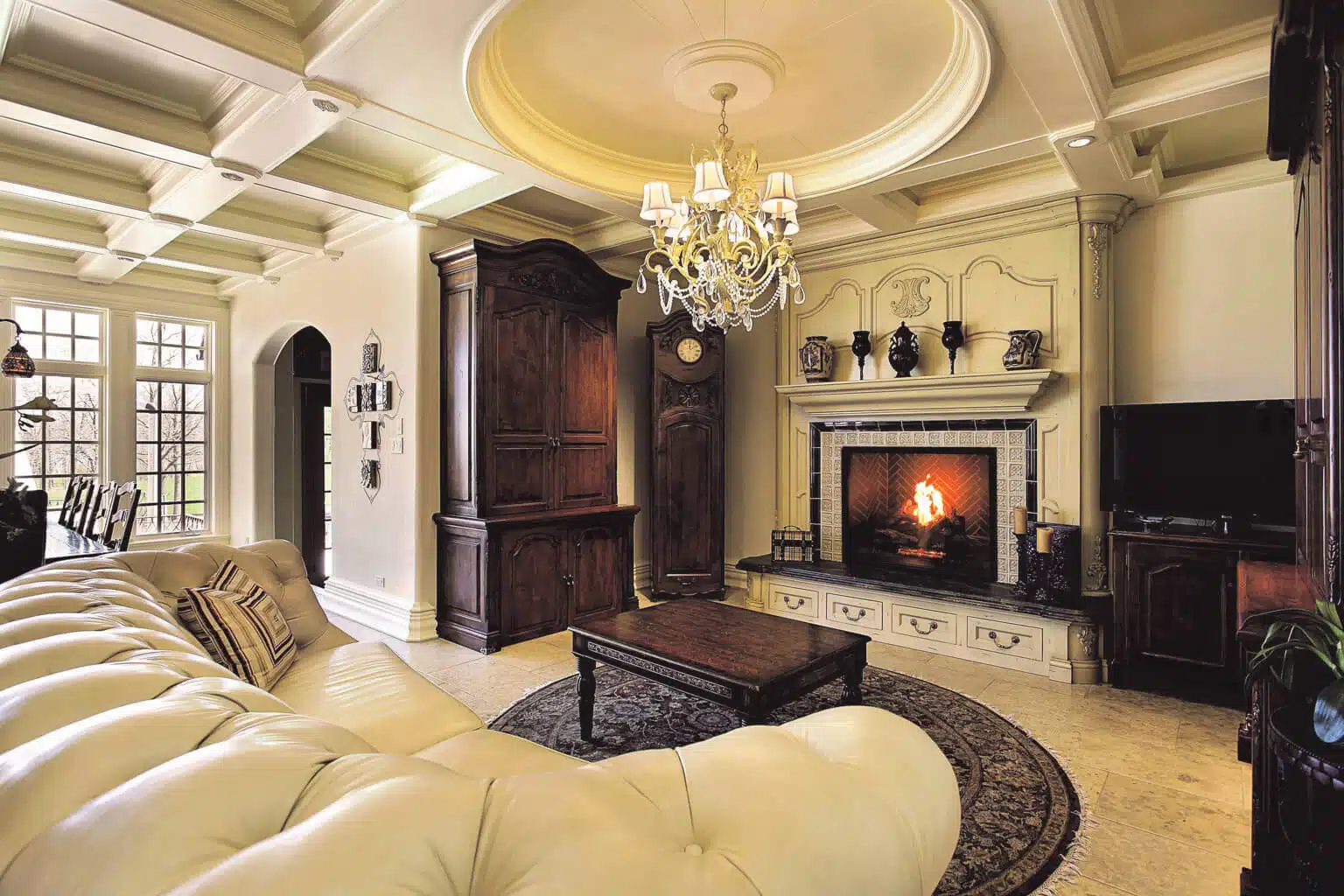 Luxurious living room featuring a white tufted sofa, ornate chandelier, and a grand fireplace with a lit fire. Dark wood furnitureluding a cabinet and coffee table, complement the elegant decor. Large glass walls allow natural light to illuminate the space beautifully.