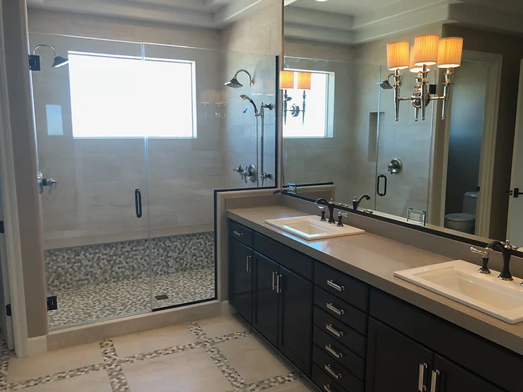 A modern bathroom featuring a double-sink vanity with black cabinets, a large mirror, and wall-mounted lights. To the left is a spacious shower area enclosed with sleek glass walls, accentuated by beige tiles and a small window. A toilet is partially visible in the corner.