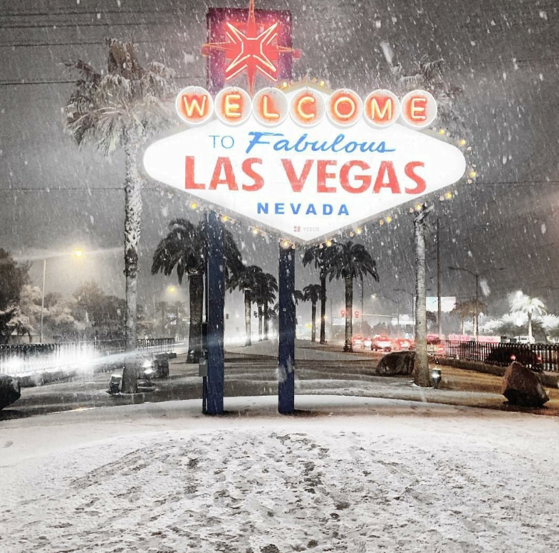 The iconic "Welcome to Fabulous Las Vegas, Nevada" sign is illuminated at night during a rare snowfall. The scene is covered in snow, with glass walls reflecting the wintry spectacle. Palm trees and traffic are visible in the background under the falling snowflakes.