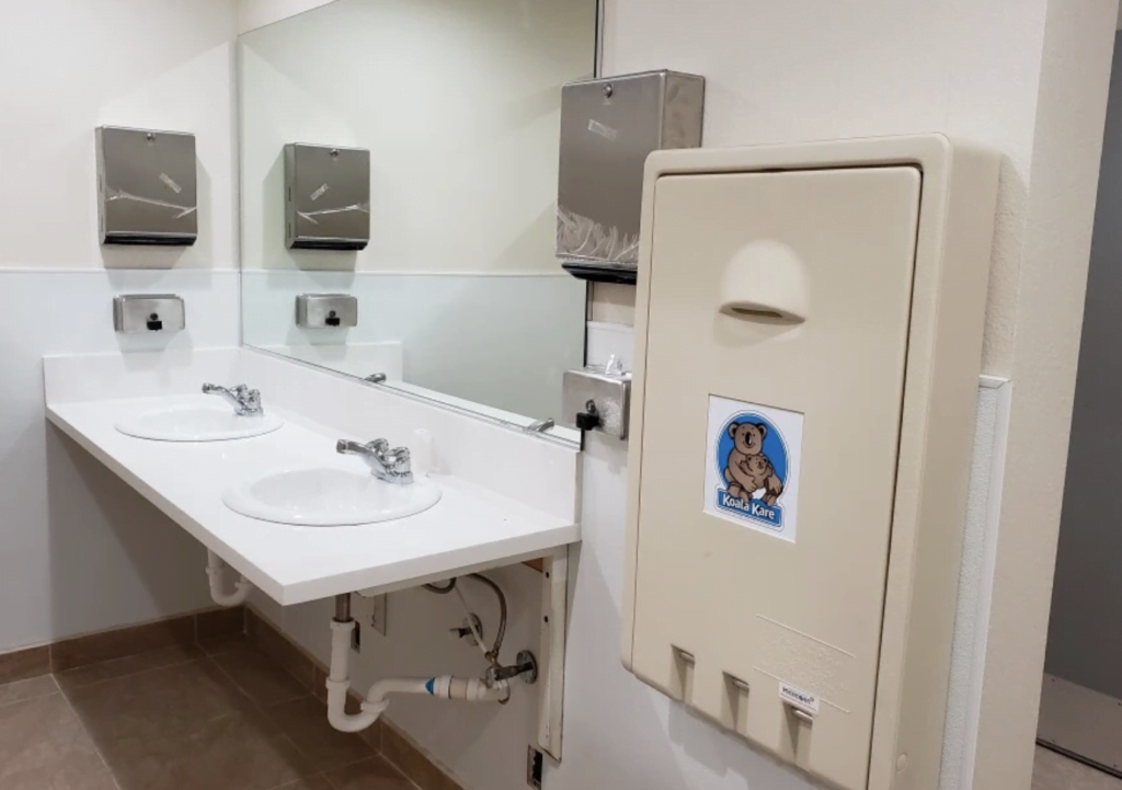 A restroom with two white sinks and mirrors above each. Two metal paper towel dispensers are mounted on the wall above the sinks. A beige baby changing station with a Koala Kare logo is affixed to the adjacent wall. The tiled floor complements the modern shower enclosures nearby.