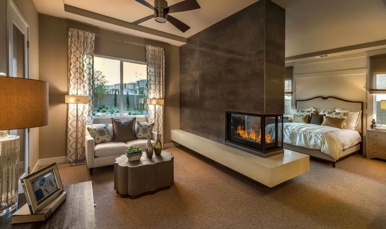 A modern bedroom with an attached sitting area, featuring a large window with drapes, a ceiling fan, and a recessed fireplace by Hearth & Home Specialties dividing the bed from the sitting area. The sitting area includes a sofa, coffee table, and decorative items.