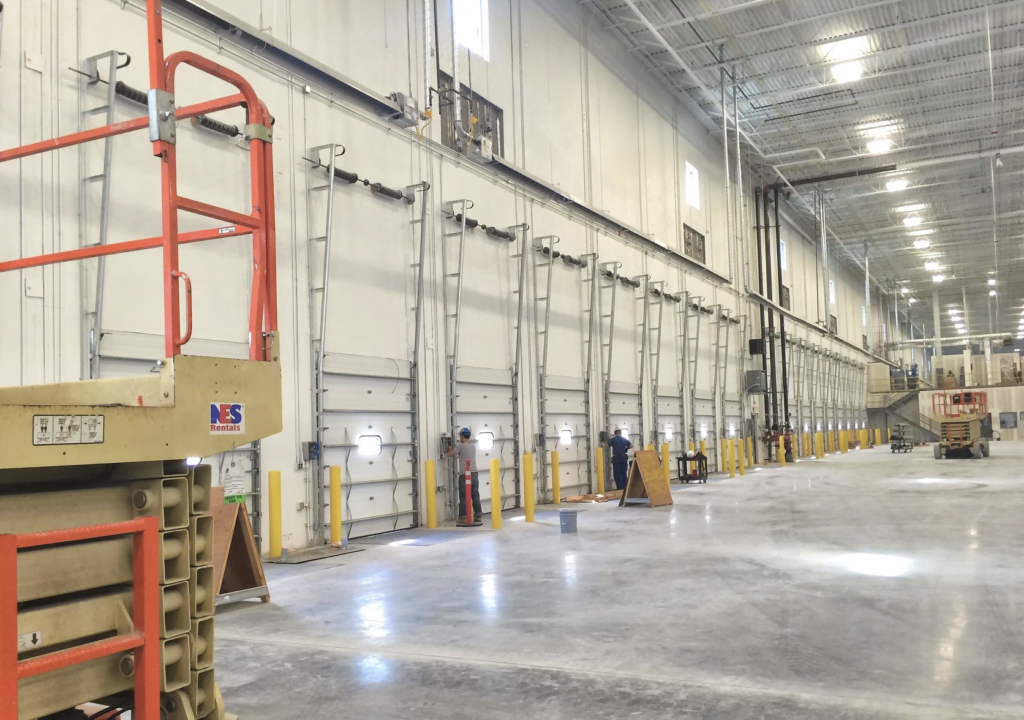 A large, spacious warehouse with high ceilings and multiple loading dock doors lined up along one wall. The facility features bright lighting and a polished concrete floor. Hearth & Home Specialties' scissor lift is in the left foreground, and a few workers are seen in the distance.