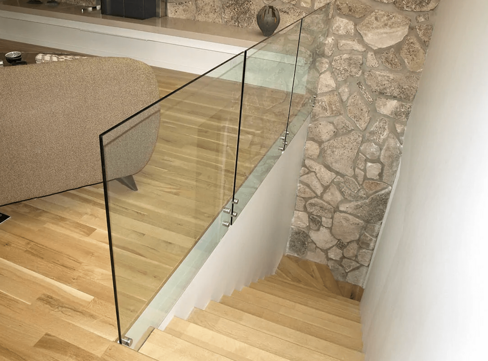 Staircase with wooden steps leading down from a living area. The staircase is enclosed by transparent glass railings with metal brackets. The living area features a stone accent wall and a brown couch on a wooden floor.