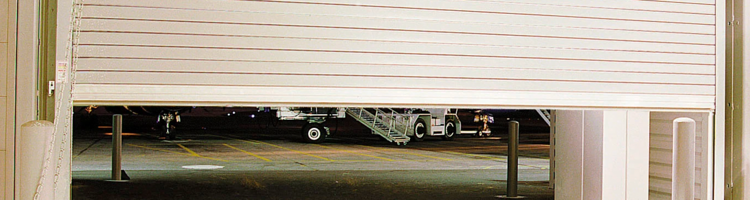 A partially open roll-up door in an industrial garage reveals an airplane parked on the tarmac outside. The scene is lit, indicating activity, with ground support equipment visible near the aircraft. The inside of the commercial garage appears dimly lit.