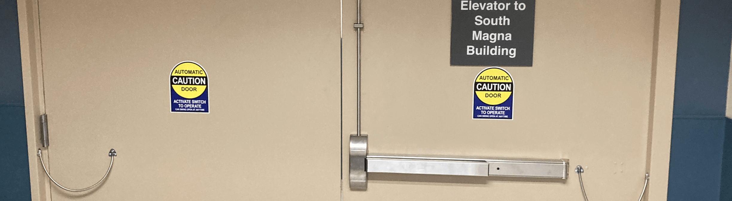 A set of beige double doors with a horizontal push bar and two caution signs indicating "Automatic Caution Door." There is a black sign above the doors reading "Elevator to South Magna Building." This commercial door installation is located in a hallway with blue and beige walls.