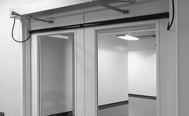 A black and white image of closed double glass fire doors set in a plain wall. The doors have windows and are framed with a dark-colored border. Above the doors is a mechanical apparatus, likely for automatic opening, with cables extending from it.