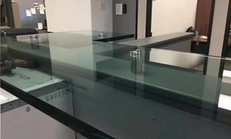 A modern office reception desk made of transparent glass with metallic supports and a sleek design. The glass countertops add an extra touch of sophistication. In the background, you can see part of another office area with some office equipment and furniture. The setting appears clean and professional.