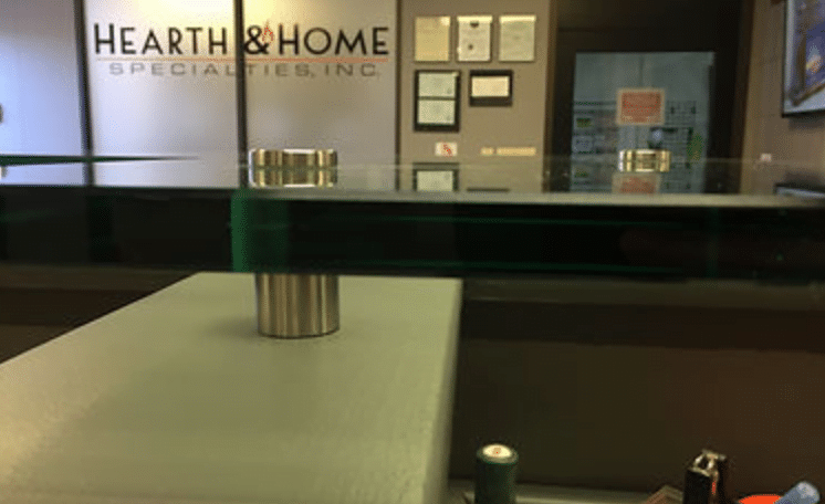 Image of a modern reception desk in an office with large glass countertops supported by metal mounts. The background features a wall with framed certificates and the sign "Hearth & Home Specialties, Inc." The area appears neat and professional.