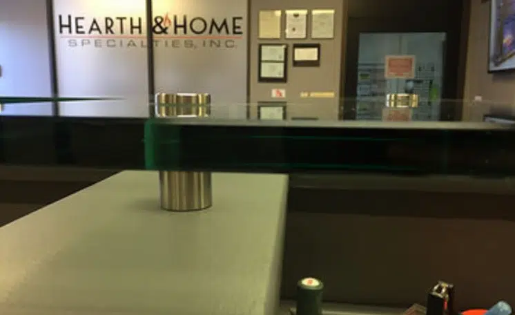 Image of a modern reception desk in an office with large glass countertops supported by metal mounts. The background features a wall with framed certificates and the sign "Hearth & Home Specialties." The area appears neat and professional.