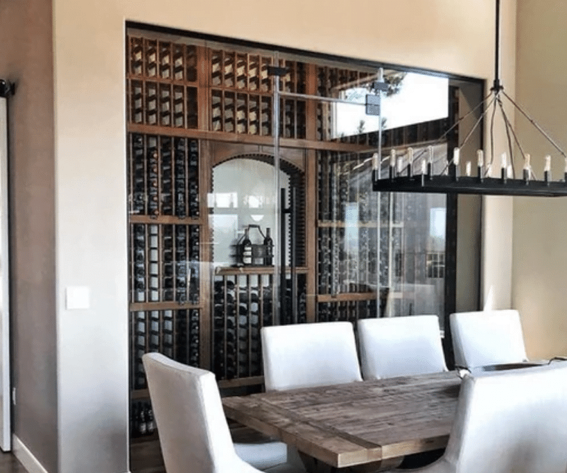 A modern dining room features a wooden table surrounded by white upholstered chairs. Behind the dining area, glass wall partitions enclose a large wine cellar, with numerous bottles neatly arranged on wooden racks. A contemporary chandelier hangs above the table, adding an elegant touch.