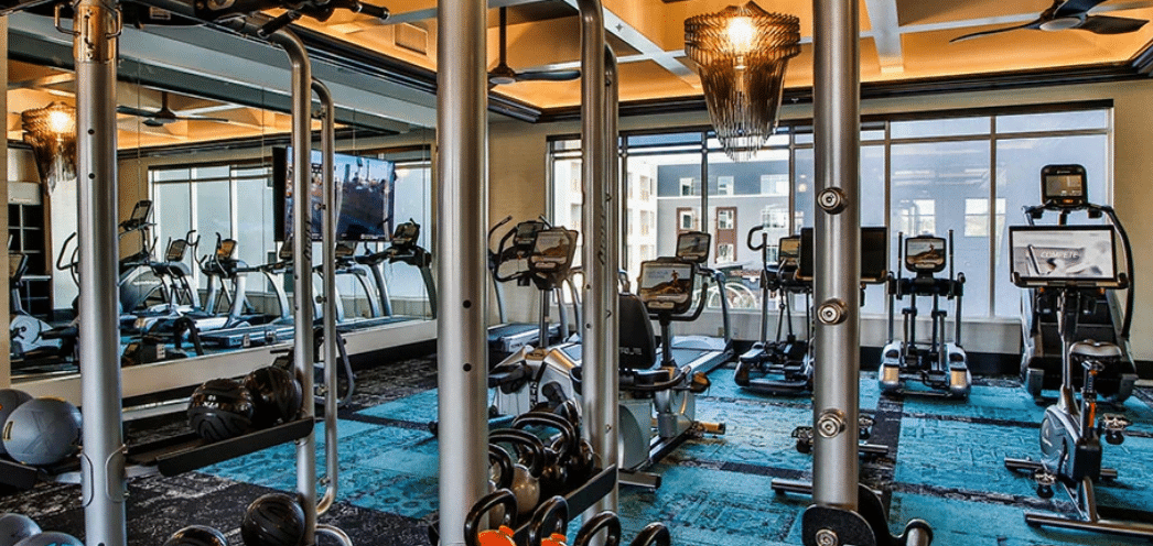 A modern gym with various exercise equipment including treadmills, stationary bikes, and weightlifting machines. The space features large wall mirrors, teal and black flooring, and ample natural light from the windows. A wall-mounted TV is turned on in the background.