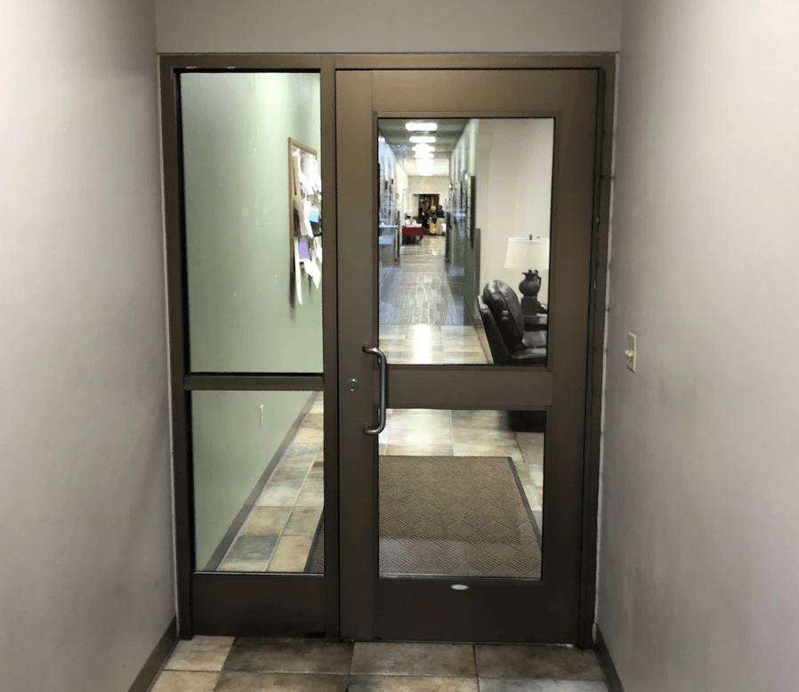 A commercial door with a metal frame is located at the end of a hallway, leading to a well-lit corridor with green walls and tile flooring. In the corridor, a bulletin board, chairs, and framed pictures are visible.