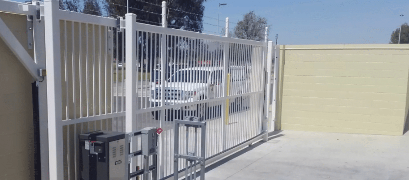 A white automated sliding gate with vertical bars is shown partially open, offering a glimpse of a white vehicle behind it. Flanked by beige walls, the entry features various control mechanisms visible on the left side.