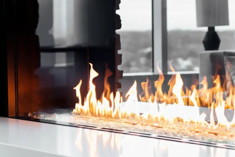 A modern linear fireplace with a wide, rectangular glass enclosure from Hearth & Home Specialties. Bright flames dance over a bed of shiny pebbles. In the background, blurred natural light filters through large windows of a contemporary living space.